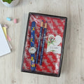 Set of pencils and a watch in a decorative box on a wooden surface with stationery items.